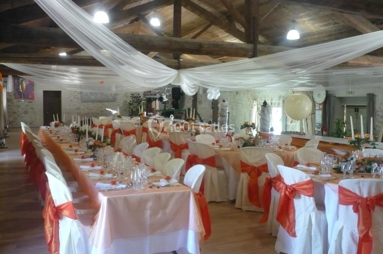 Salle de réception décorée pour un événement, avec tables alignées, nappes blanches et nœuds orange sur les chaises.