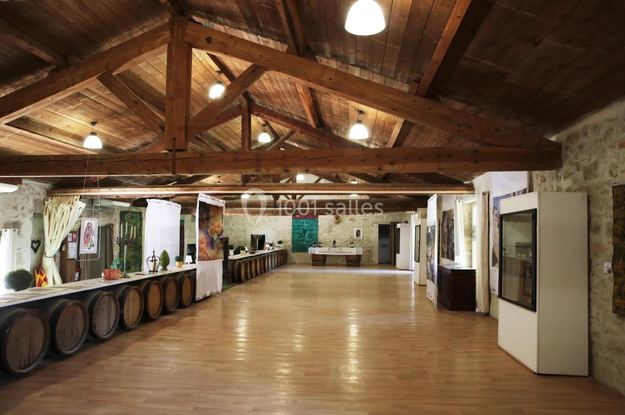 Salle en pierre avec plafond en poutres apparentes, barriques en bois alignées et éclairage suspendu.