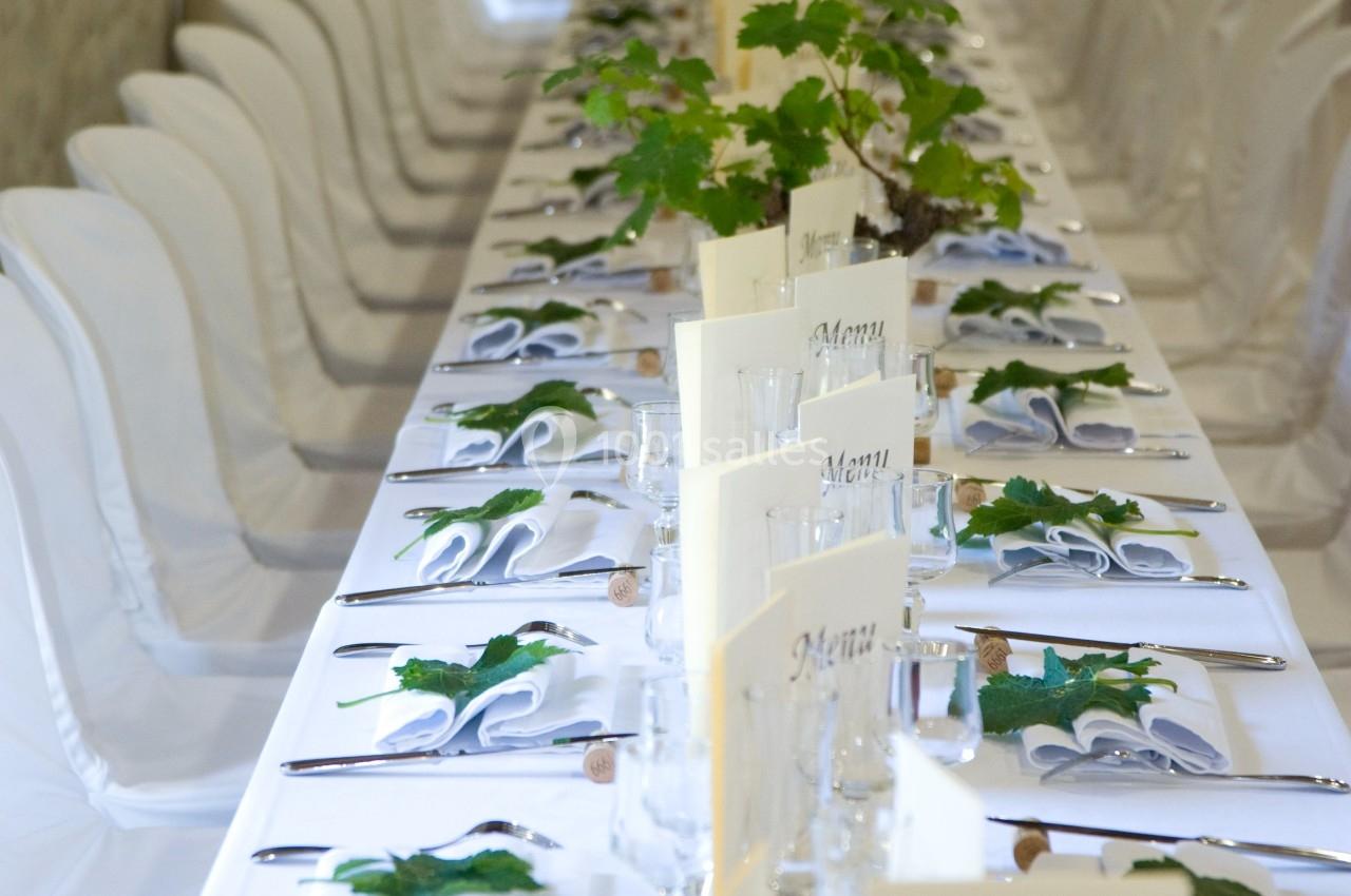 Table de banquet élégamment dressée avec nappes blanches, menus, couverts alignés et décoration de feuilles vertes.