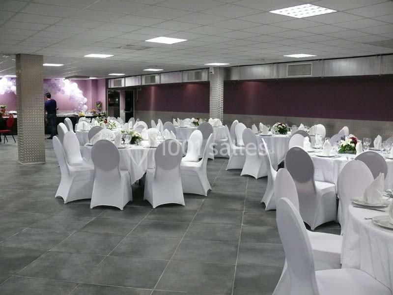 Salle de réception avec tables rondes dressées, nappes blanches et chaises houssées, éclairée par un plafond lumineux.