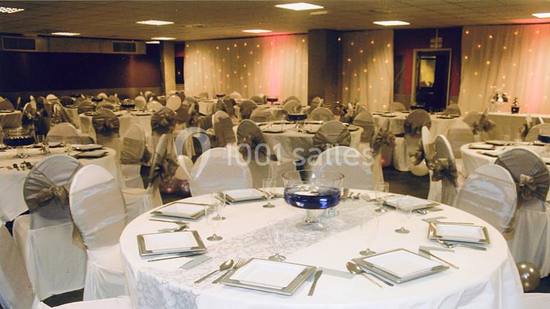 Salle de réception décorée avec des tables rondes dressées, nappes blanches et chaises ornées de nœuds.