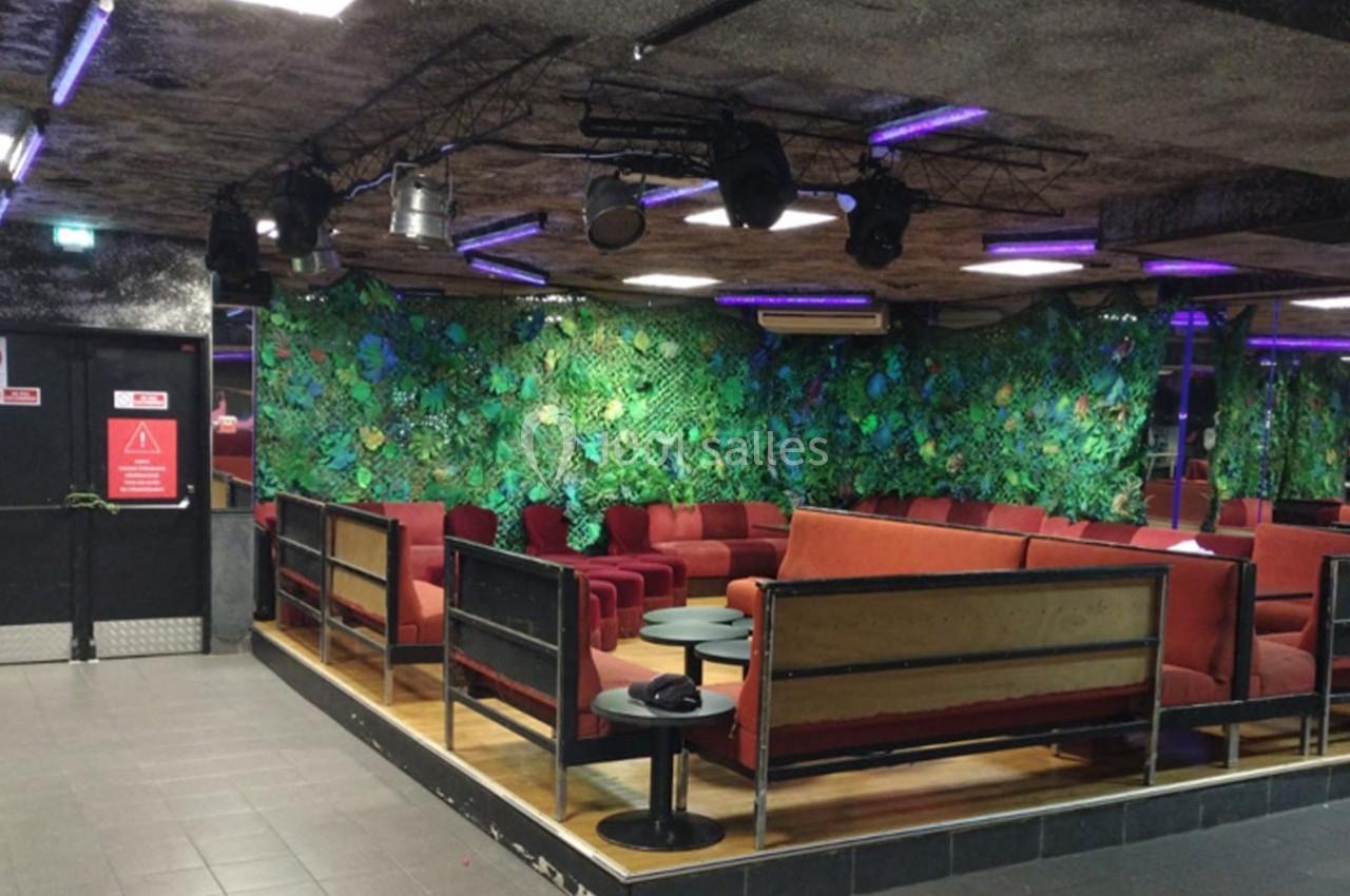 Espace intérieur avec banquettes rouges, tables basses et murs décorés de feuillage artificiel sous éclairage violet.