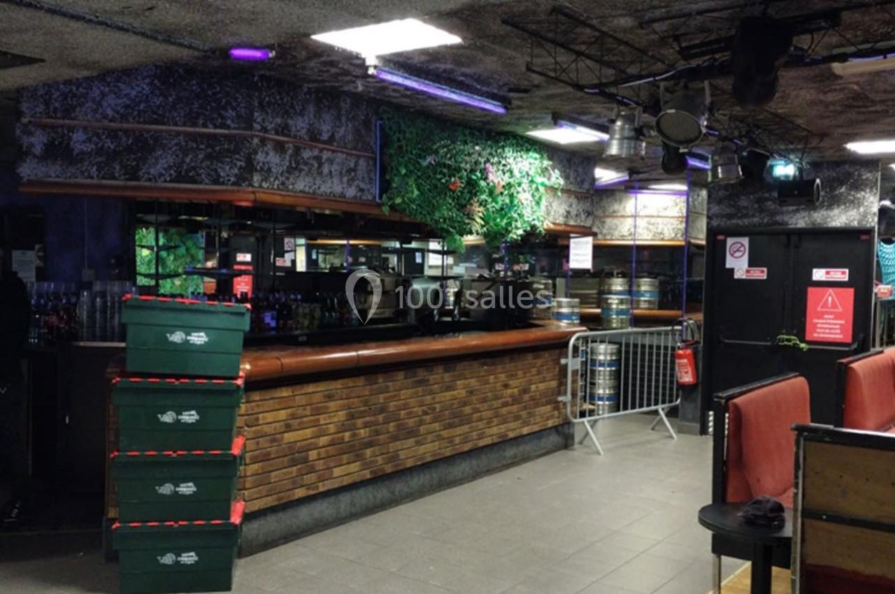 Bar intérieur avec comptoir en briques, caisses empilées, fûts de bière, et décor végétal en arrière-plan.