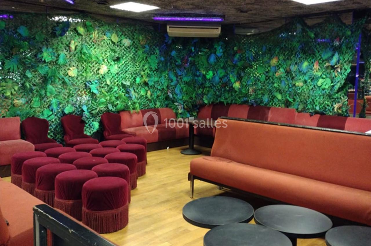 Salle avec banquettes rouges, tabourets assortis, tables basses noires et mur décoré de végétation artificielle.