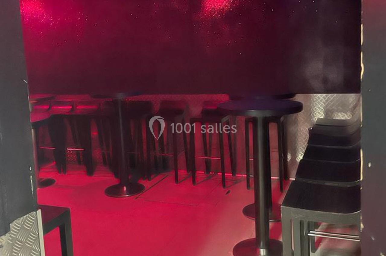 Salle sombre avec éclairage rouge, tables hautes noires et tabourets alignés le long des murs métalliques.