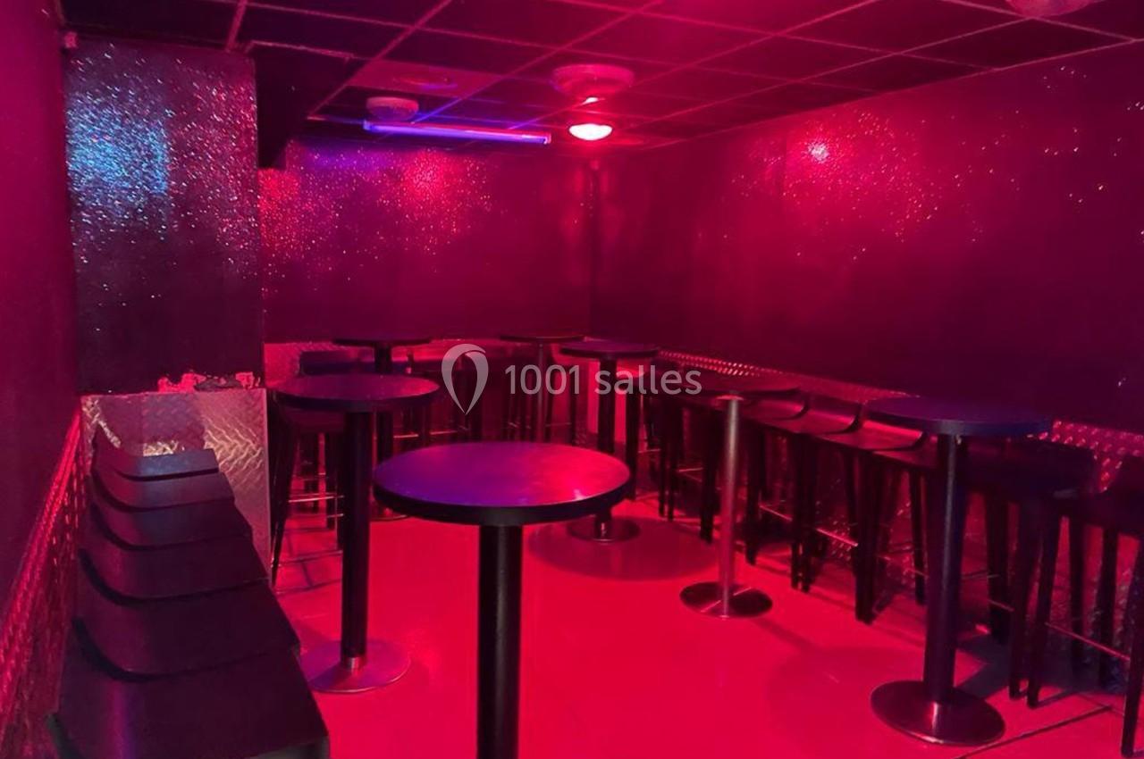 Salle sombre avec éclairage rouge, tables hautes noires, tabourets alignés et murs scintillants.