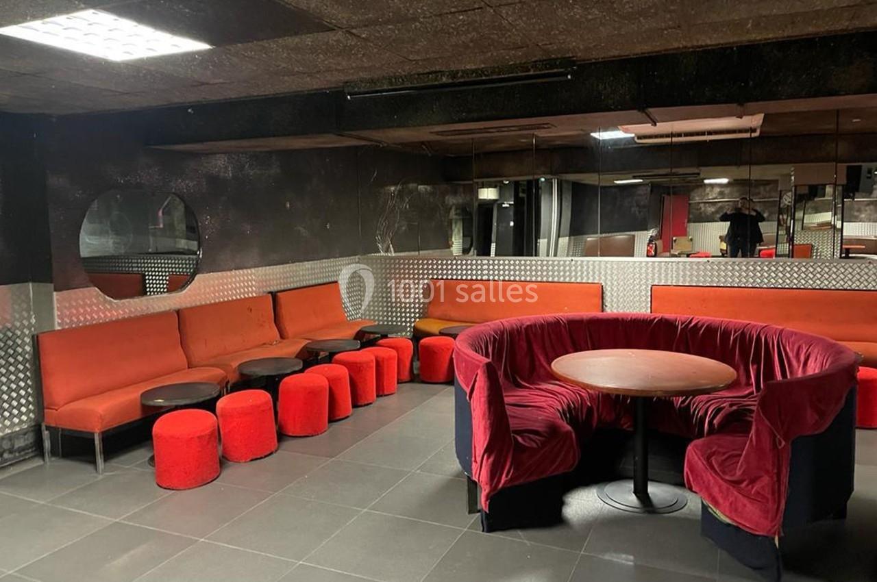 Salle avec mobilier rouge et noir, comprenant des banquettes, poufs et une table ronde, éclairée par des néons au plafond.