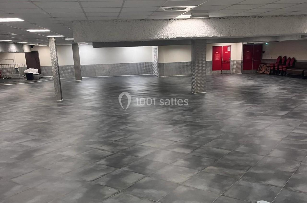 Salle vide avec sol en carrelage gris, piliers métalliques et murs clairs, éclairée par un plafond à dalles.