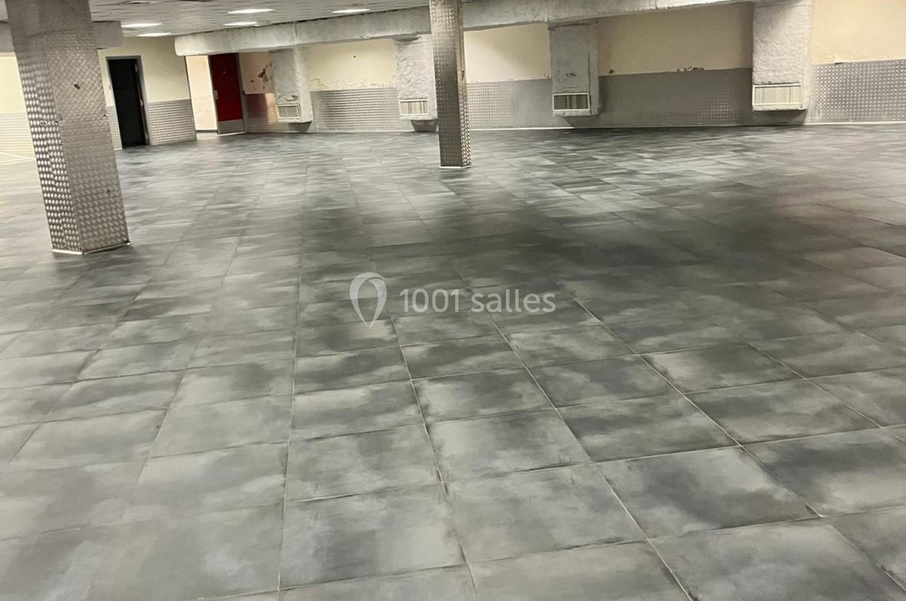 Salle vide avec un sol en carrelage gris, colonnes recouvertes de mosaïques et murs clairs partiellement carrelés.