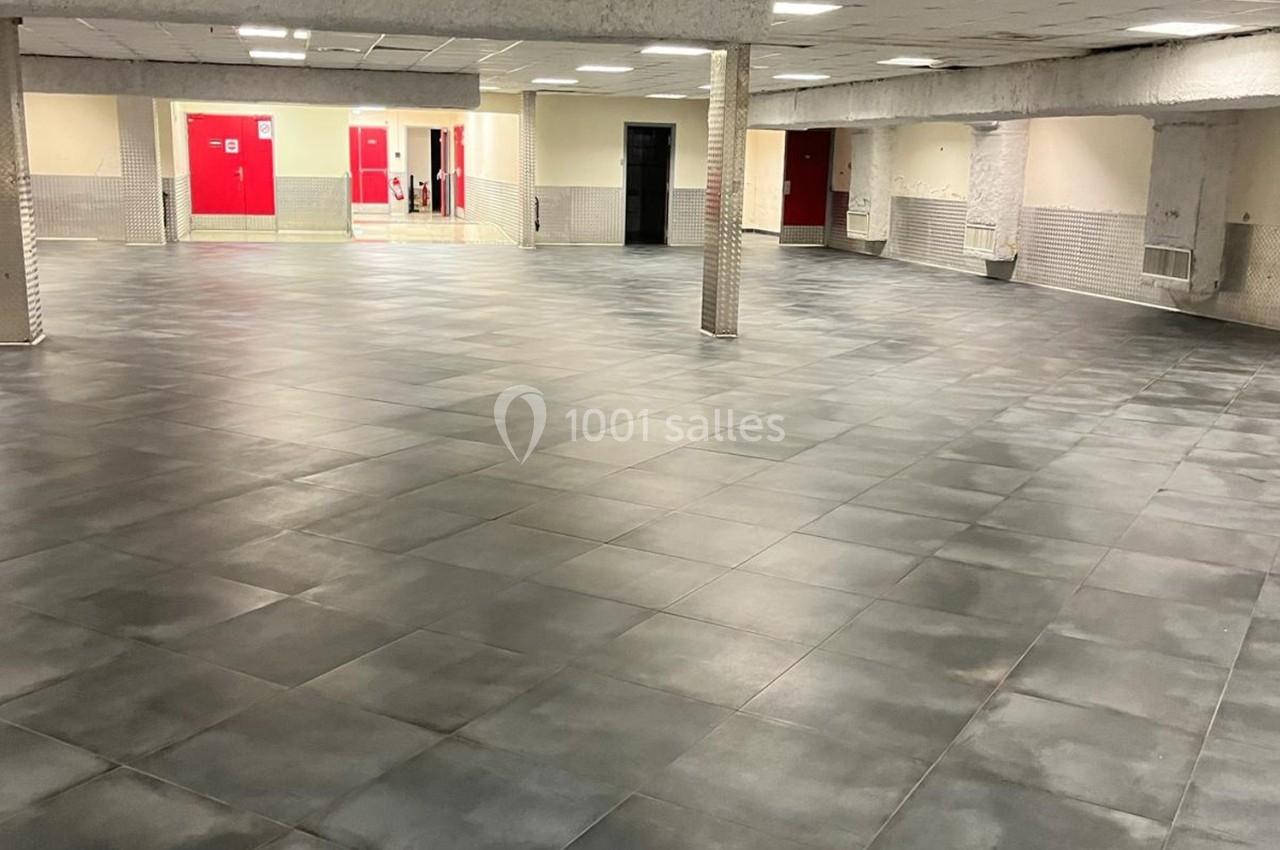 Salle vide avec un sol en carrelage gris foncé, des murs clairs et des portes rouges visibles en arrière-plan.