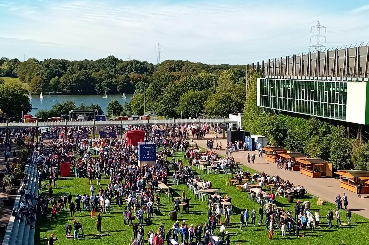 Foule rassemblée dans un espace vert avec des stands, près d'un bâtiment moderne et d'un lac bordé d'arbres.