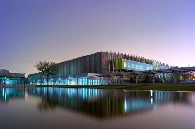 Bâtiment moderne avec façade vitrée, éclairé au crépuscule, reflété dans un plan d'eau calme.