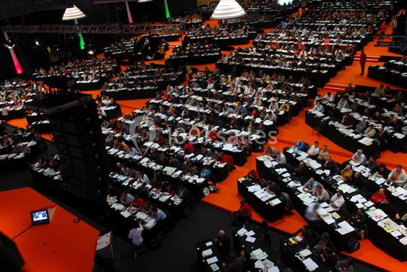 Salle de conférence bondée avec de nombreuses personnes assises à des tables, éclairage tamisé et ambiance professionnelle.