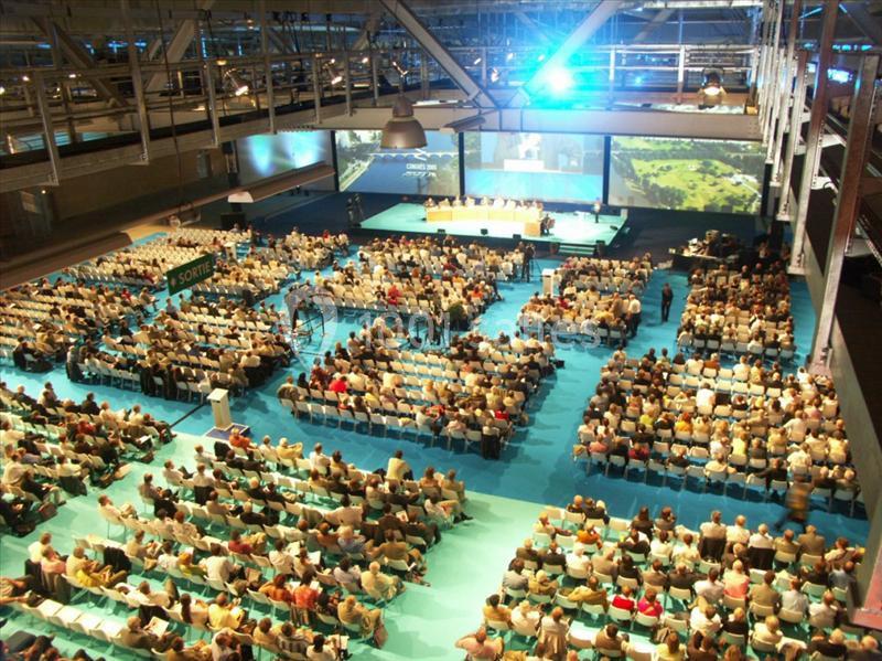Salle de conférence bondée avec des participants assis, une scène principale et plusieurs écrans de projection.