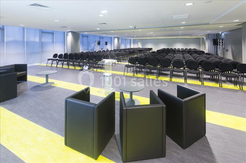 Salle de conférence vide avec rangées de chaises noires, estrade en plexiglas et fauteuils modernes au premier plan.