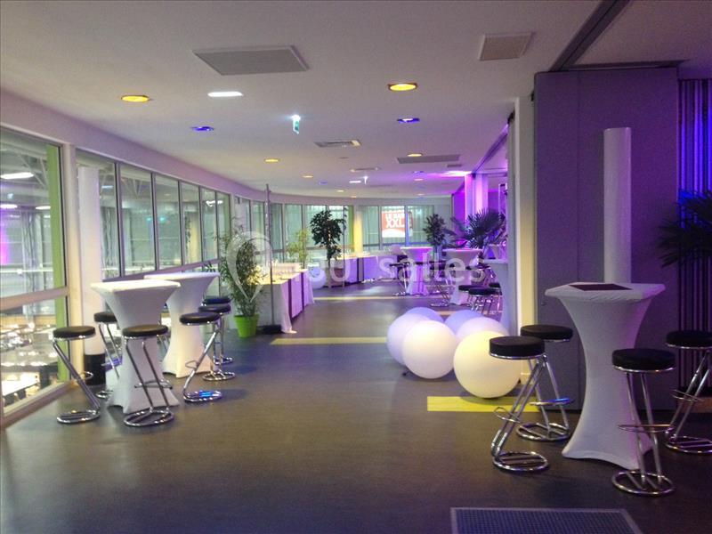 Salle lumineuse avec tables hautes, tabourets noirs, plantes et éclairage coloré, donnant sur des baies vitrées.