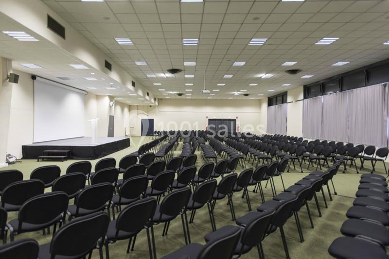 Salle de conférence vide avec des rangées de chaises noires alignées et une estrade équipée d'un pupitre.