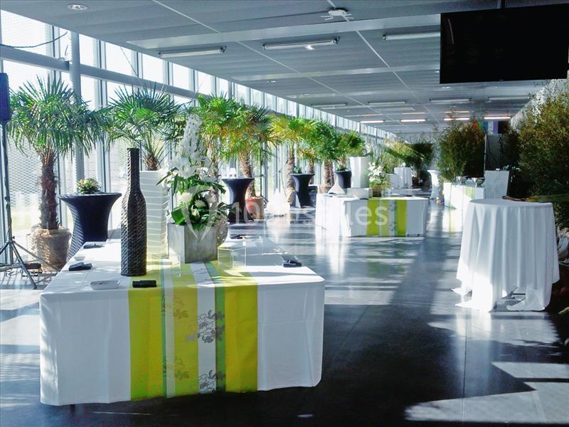 Salle lumineuse avec grandes baies vitrées, tables décorées de nappes blanches et vertes, plantes en arrière-plan.