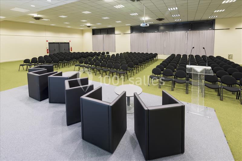 Salle de conférence avec chaises alignées, scène équipée de fauteuils, table basse et pupitre transparent.
