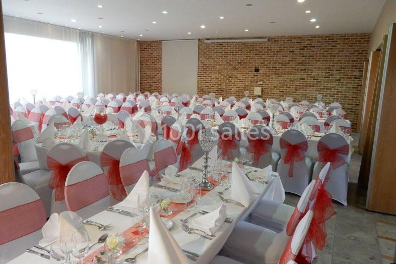 Salle de réception décorée avec des nappes blanches et des chaises ornées de nœuds rouges, tables dressées pour un événement.