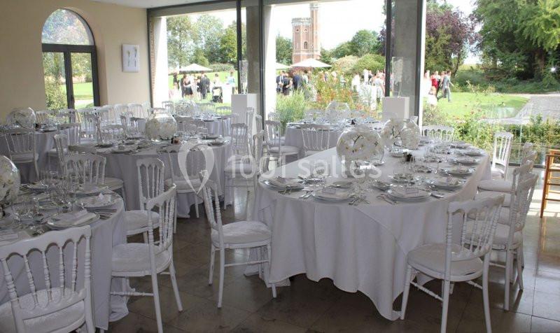 Salle de réception lumineuse avec tables rondes et ovales dressées, vue sur un jardin et des invités à l'extérieur.