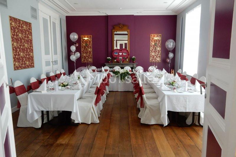 Salle décorée pour un événement avec tables dressées, nappes blanches, chaises ornées de rubans rouges et ballons argentés.