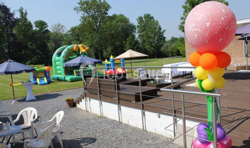 Espace extérieur avec structures gonflables, terrasse en bois, tables et chaises sous parasols, décoré de ballons colorés.