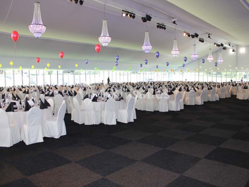 Salle de réception spacieuse avec des tables dressées, des chaises blanches et des ballons colorés suspendus.