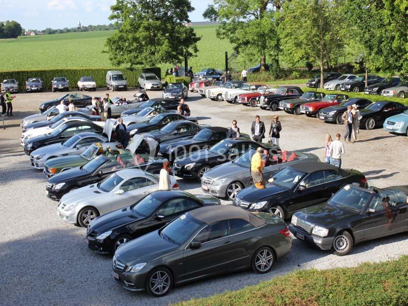 Rassemblement de voitures de luxe sur un parking en plein air, entouré de verdure et de participants.