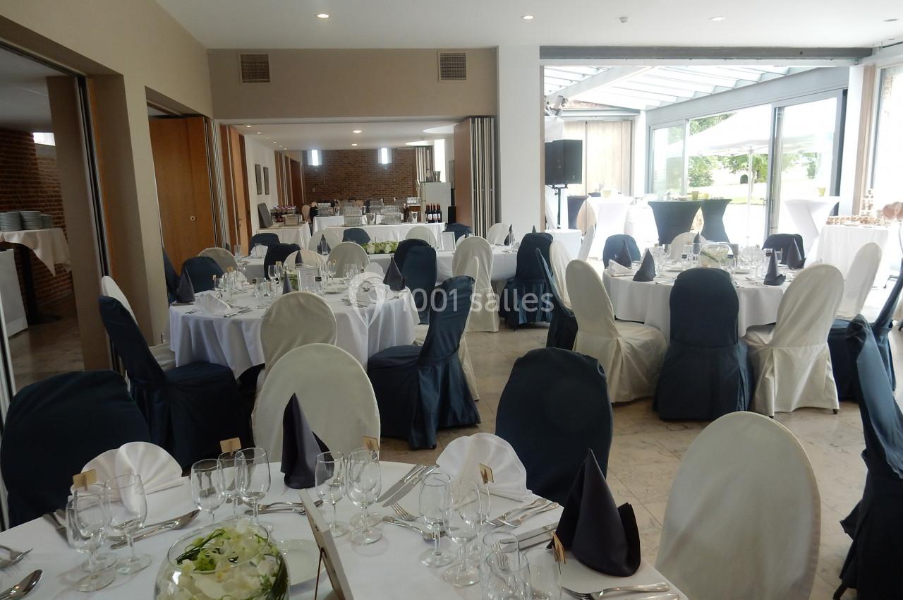 Salle de réception lumineuse avec tables rondes dressées, nappes blanches et chaises couvertes de housses noires et blanches.