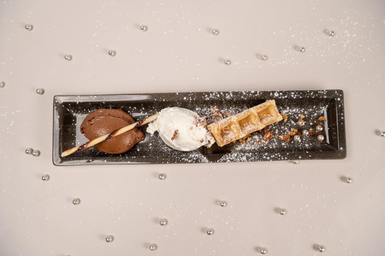 Assiette rectangulaire avec boule de glace au chocolat, boule de glace vanille, gaufre, éclats de noisettes et sucre glace.
