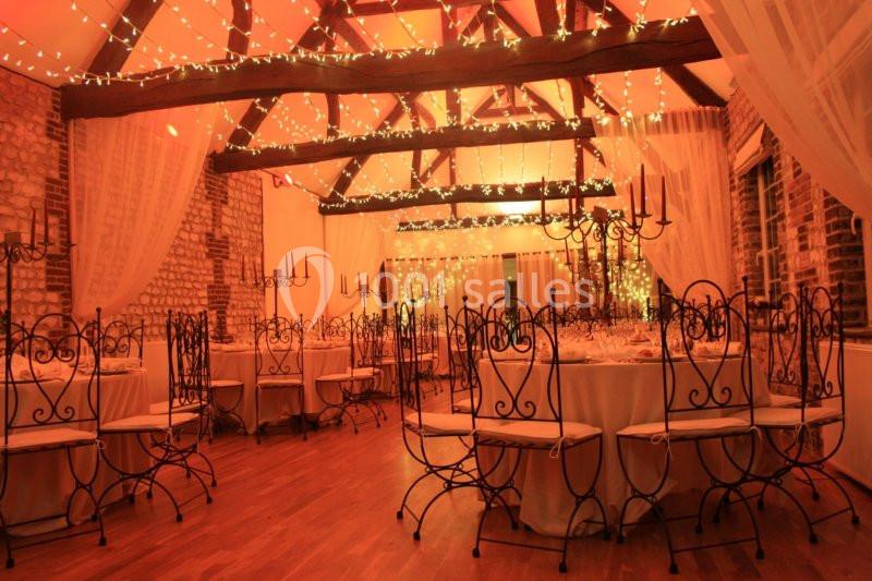 Salle de réception chaleureuse avec tables rondes, chaises en fer forgé et guirlandes lumineuses suspendues.