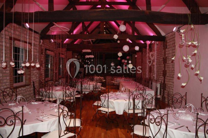Salle de réception décorée avec des tables dressées, des chaises en fer forgé et un éclairage rose sous des poutres…