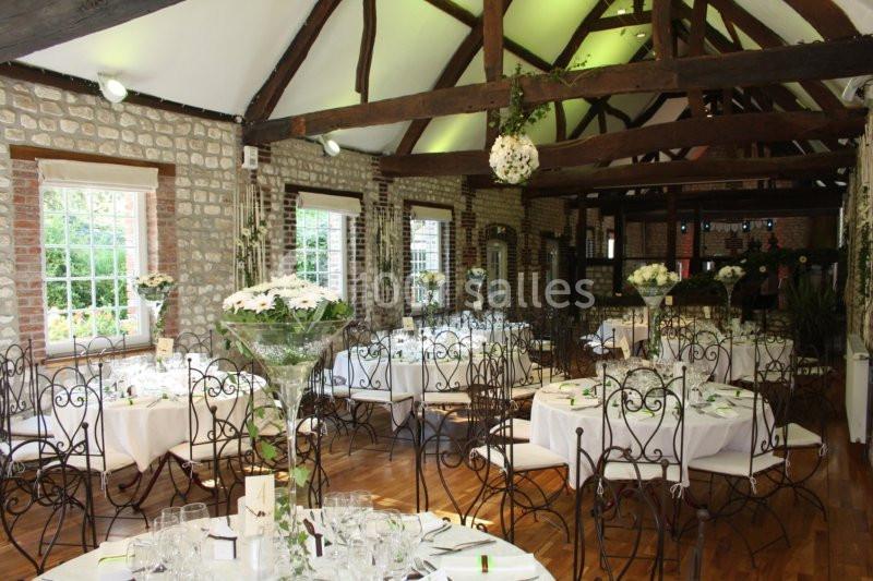 Salle de réception avec murs en pierre, poutres apparentes, tables décorées de fleurs et chaises en fer forgé.