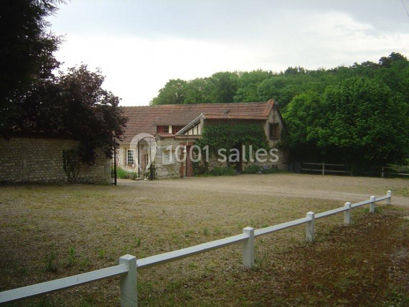 Cour gravillonnée devant un bâtiment en pierre avec toit en tuiles, entouré de végétation et d'une clôture blanche.