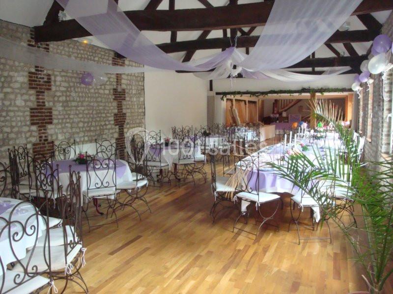 Salle de réception décorée avec des tables dressées, des nappes blanches et violettes, et des guirlandes suspendues.