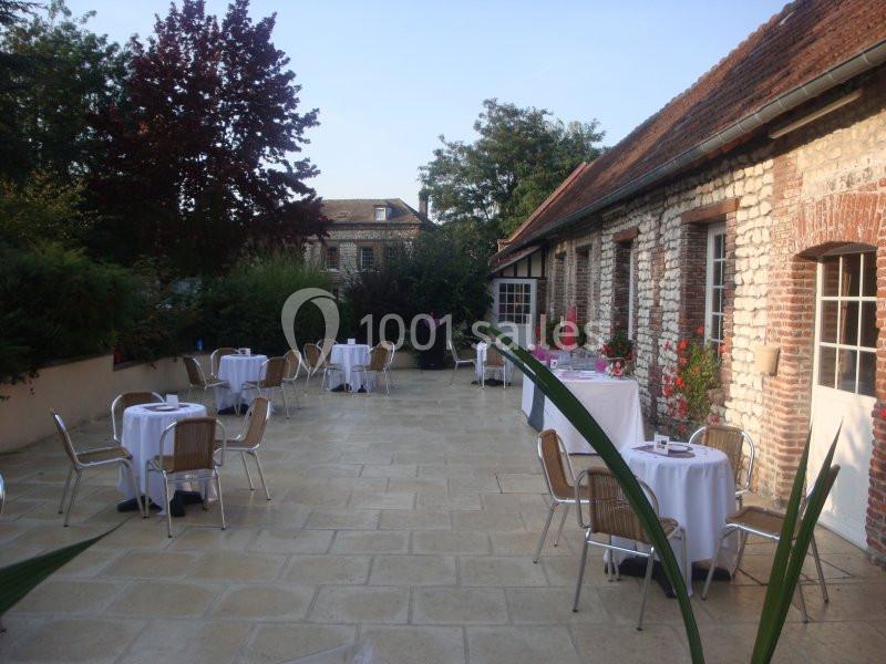 Terrasse pavée avec tables rondes dressées, entourée de bâtiments en pierre et de végétation.