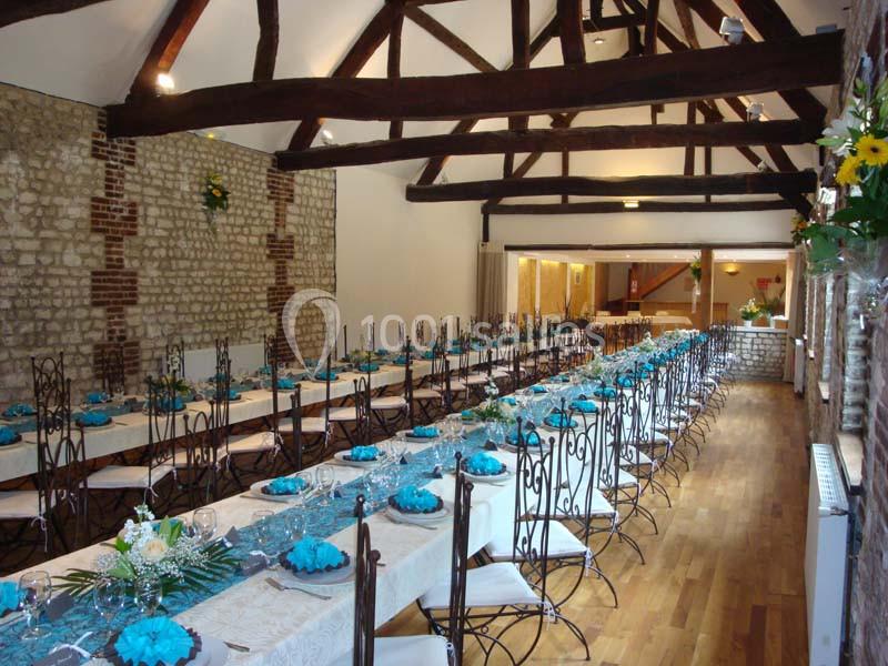 Salle de réception avec de longues tables dressées, nappes blanches et décorations bleues, sous une charpente en bois…