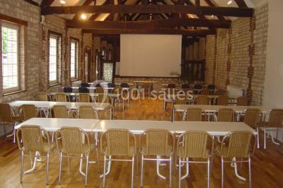 Salle de réunion avec des tables blanches, des chaises en osier et un écran de projection, dans un cadre en briques…
