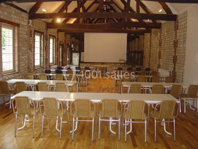Salle de réunion avec des tables blanches, des chaises en osier et un écran de projection, dans un cadre en briques…