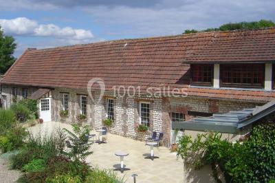 Salle de réunion avec des tables blanches, des chaises en osier et un écran de projection, dans un cadre en briques…
