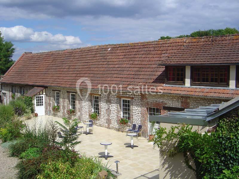 Bâtiment en pierre avec toit en tuiles, fenêtres fleuries et terrasse aménagée avec tables et chaises.