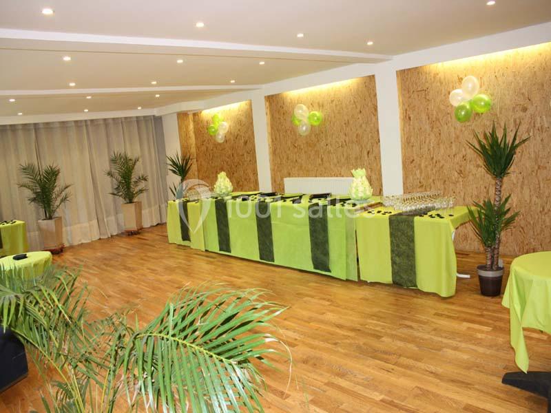 Salle décorée pour un événement avec nappes vertes, ballons assortis et plantes en pot sur un sol en parquet.