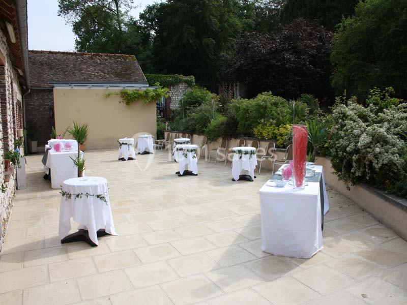 Terrasse aménagée avec des tables décorées de nappes blanches et des chaises, entourée de verdure et de bâtiments.