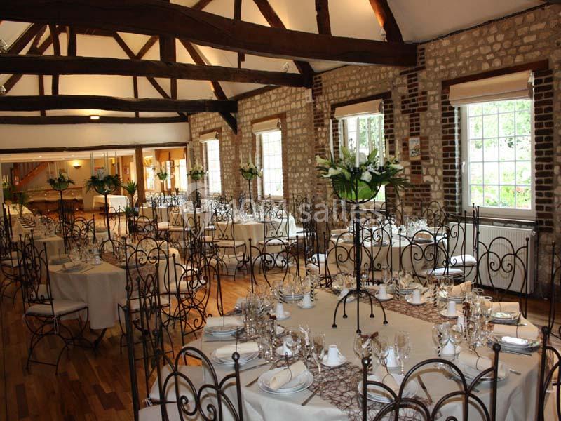 Salle de réception avec tables dressées, chaises en fer forgé, poutres apparentes et grandes fenêtres laissant entrer la…