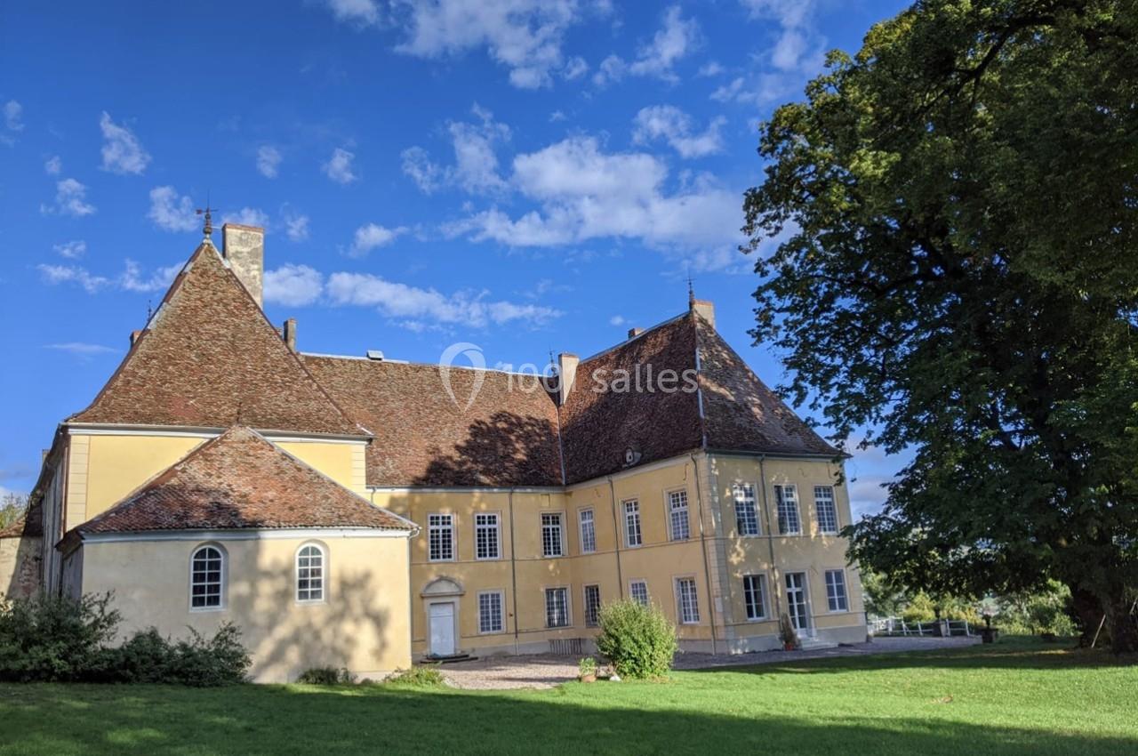 Façade d'un bâtiment historique jaune avec toit en tuiles rouges, entouré de verdure et sous un ciel bleu.