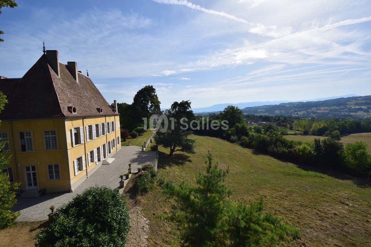 Vue d'un grand bâtiment jaune avec toit en pente, entouré de verdure et donnant sur un paysage vallonné sous un ciel dégagé.