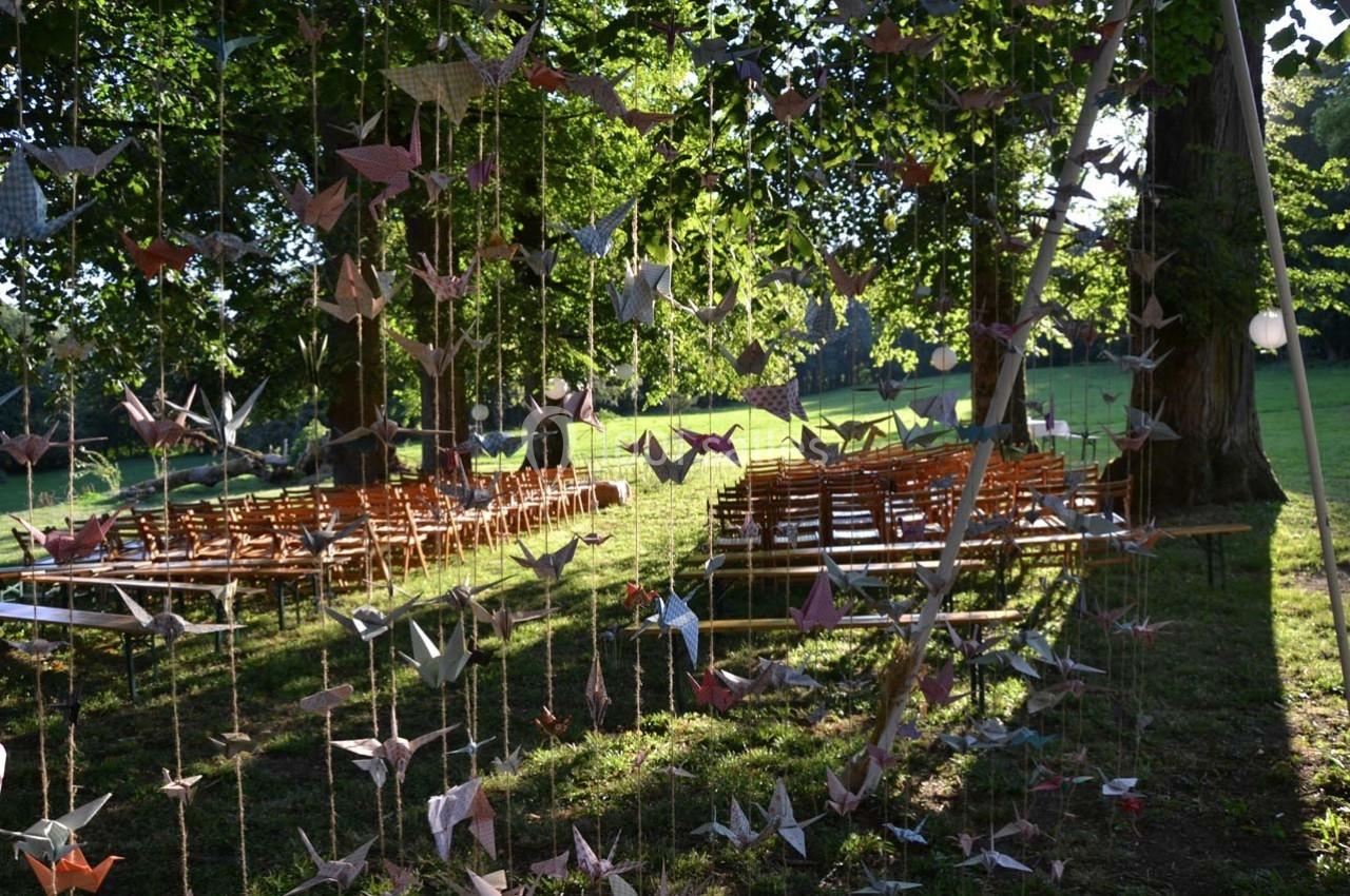 Guirlandes de grues en origami suspendues devant des rangées de chaises en bois disposées dans un parc verdoyant.