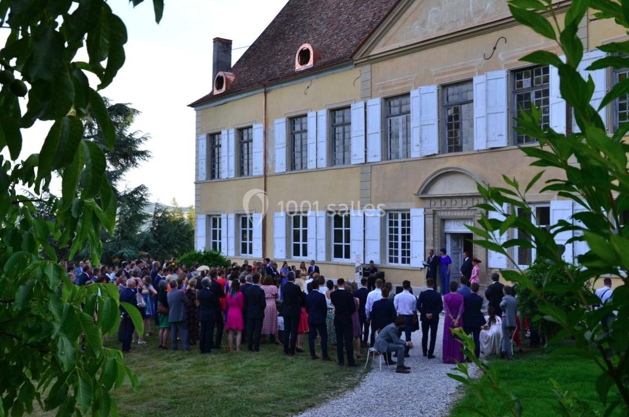 Groupe de personnes rassemblées devant une grande maison ancienne avec des volets blancs, entourée de verdure.