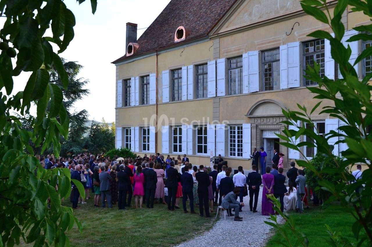 Groupe de personnes rassemblées devant une grande maison ancienne avec des volets blancs, entourée de verdure.